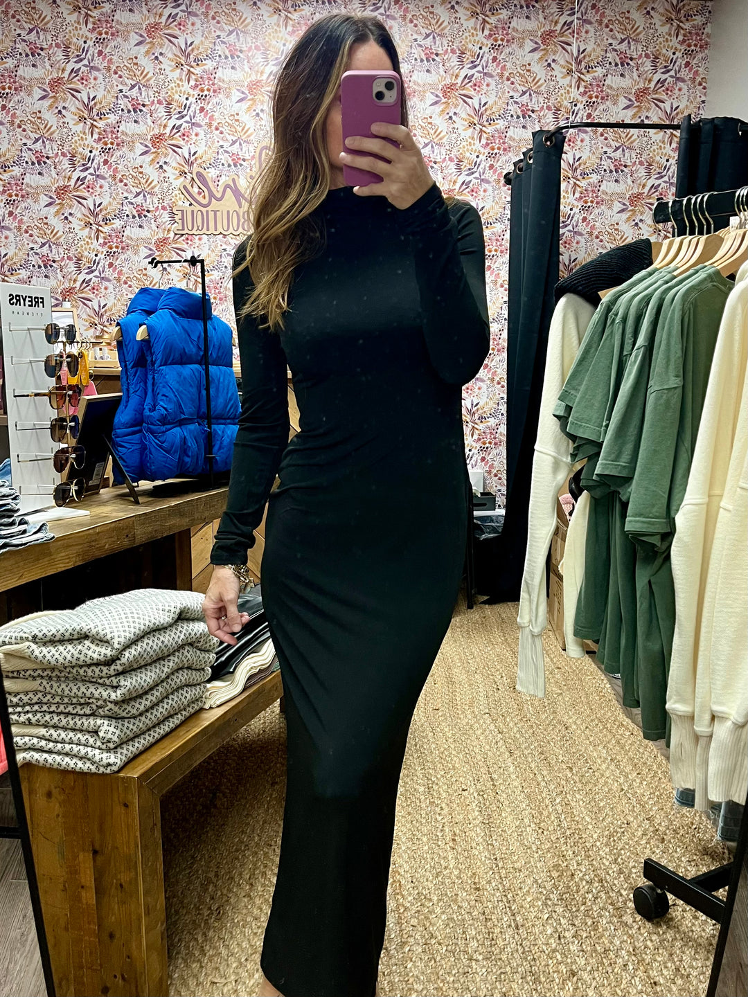 A model wearing a black turtleneck bodycon maxi dress with long sleeves, taking a picture of herself with a smartphone. The dress is displayed in a retail setting with various clothes in the background.