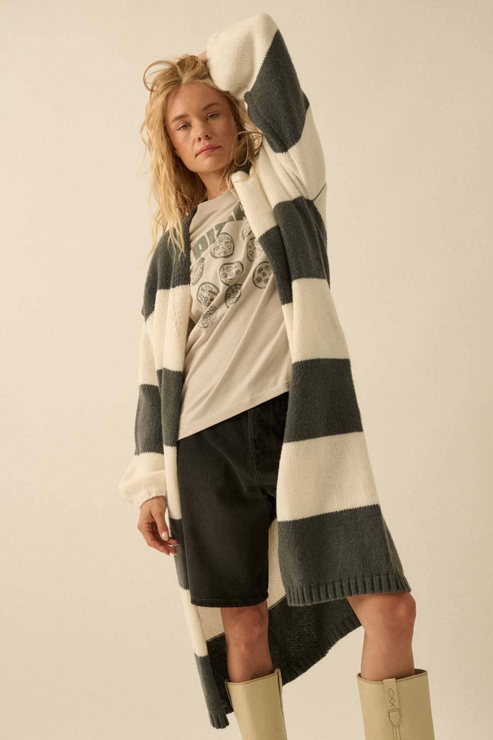 Woman wearing a long charcoal gray and off white striped cardigan over a graphic t-shirt and shorts, standing against a plain background.