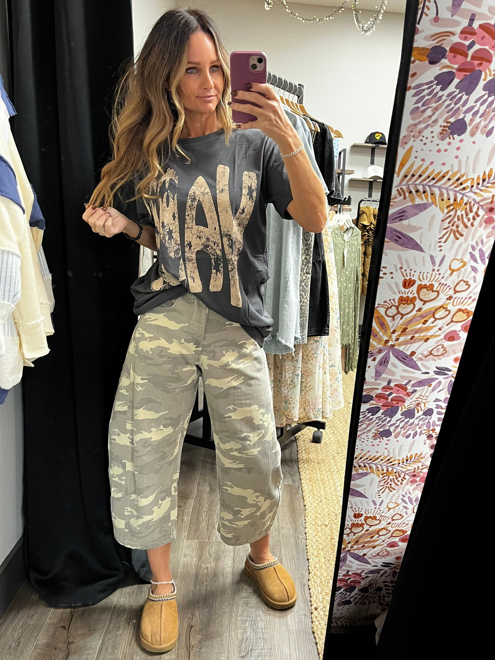 Person taking a mirror selfie wearing a graphic t-shirt and camouflage pants in a room with floral wallpaper.