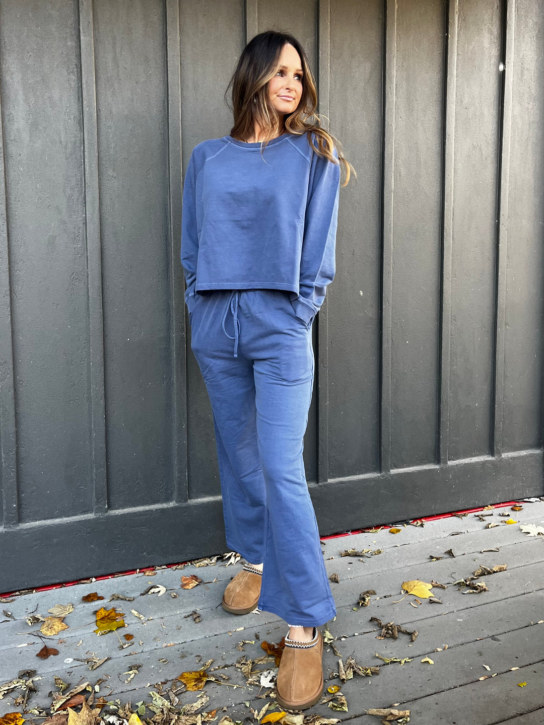 Woman wearing a navy blue outfit French terry lounge set standing against a gray wooden wall with fallen leaves on the ground.