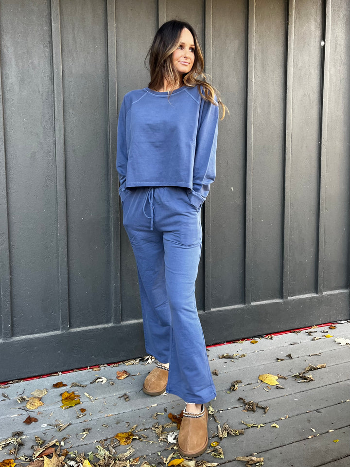 Woman wearing a navy blue outfit French terry lounge set standing against a gray wooden wall with fallen leaves on the ground.
