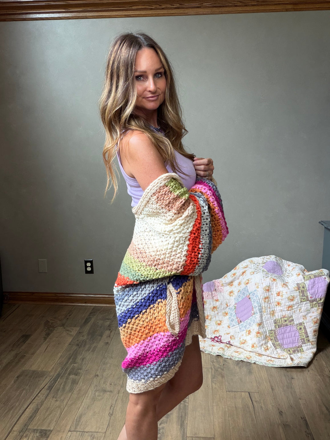 A woman stands indoors holding a chunky crocheted cardigan wrapped around her body, showing its multicolor pattern. The cardigan features a rainbow of colors including pink, orange, yellow, green, blue, and purple.
