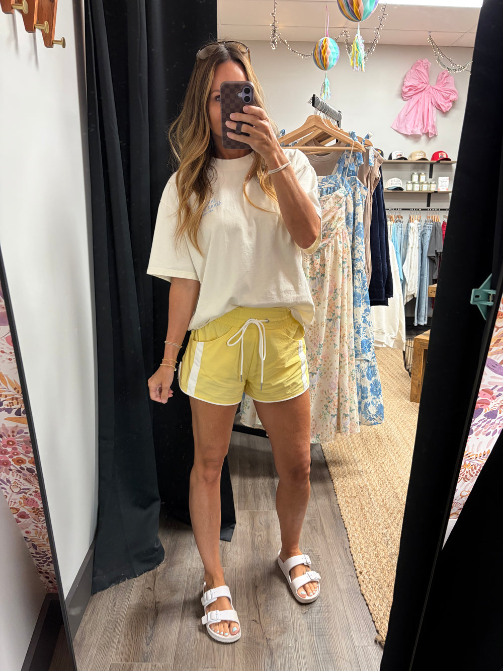 A woman takes a mirror selfie in a boutique fitting room, wearing a cream-colored oversized t-shirt, bright yellow athletic shorts with white side stripes, and white slide sandals.