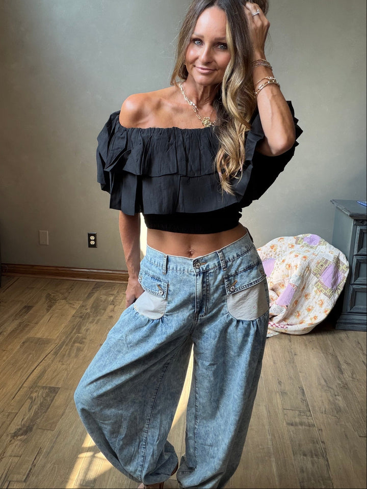 A woman wearing a black off-the-shoulder ruffled crop top and light-wash wide-leg denim pants with patch pockets.