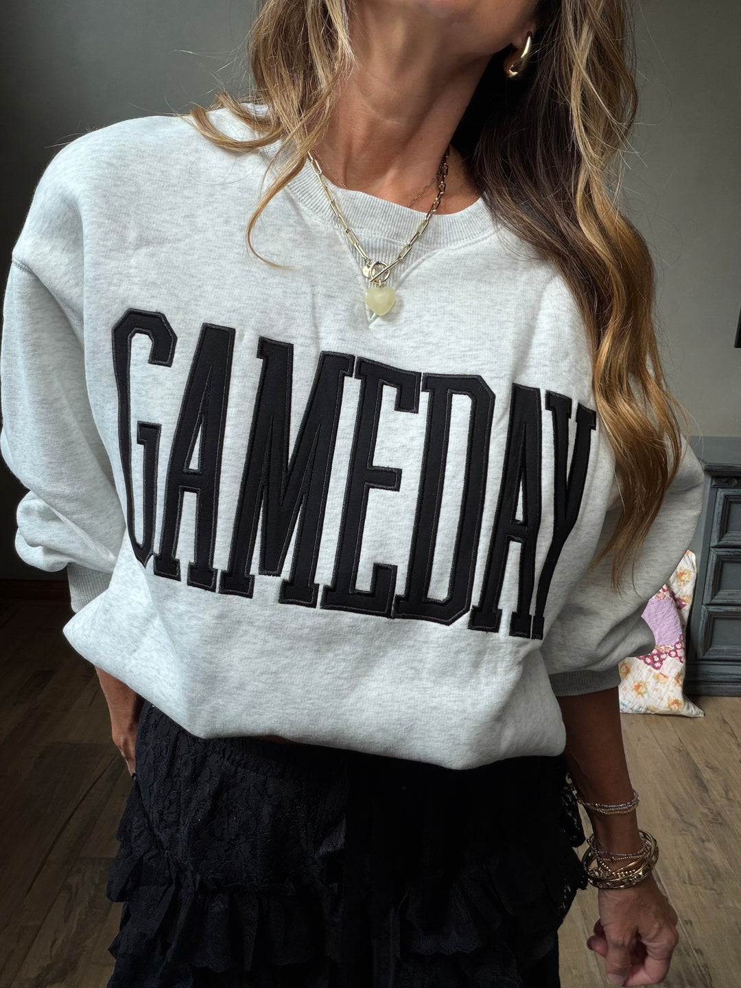 A woman wearing a light heather gray sweatshirt with the word 'GAMEDAY' printed across the front in large black letters, paired with a black ruffled skirt.