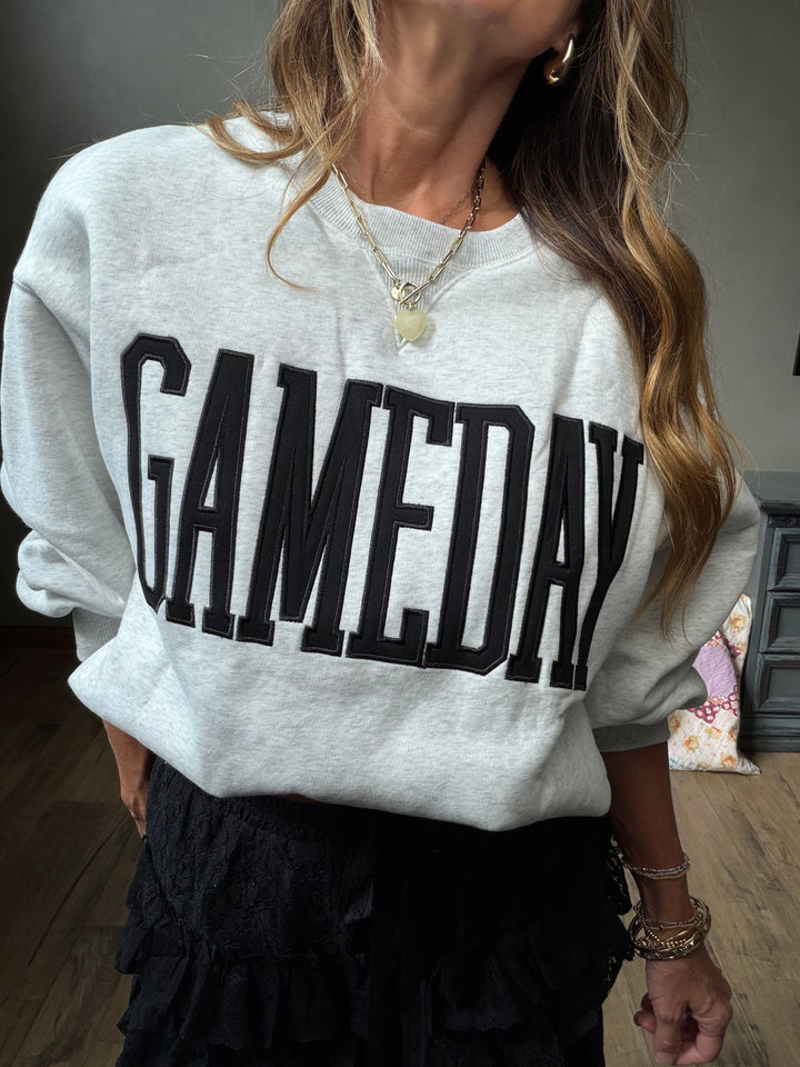 A woman wearing a light heather gray sweatshirt with the word 'GAMEDAY' printed across the front in large black letters, paired with a black ruffled skirt.